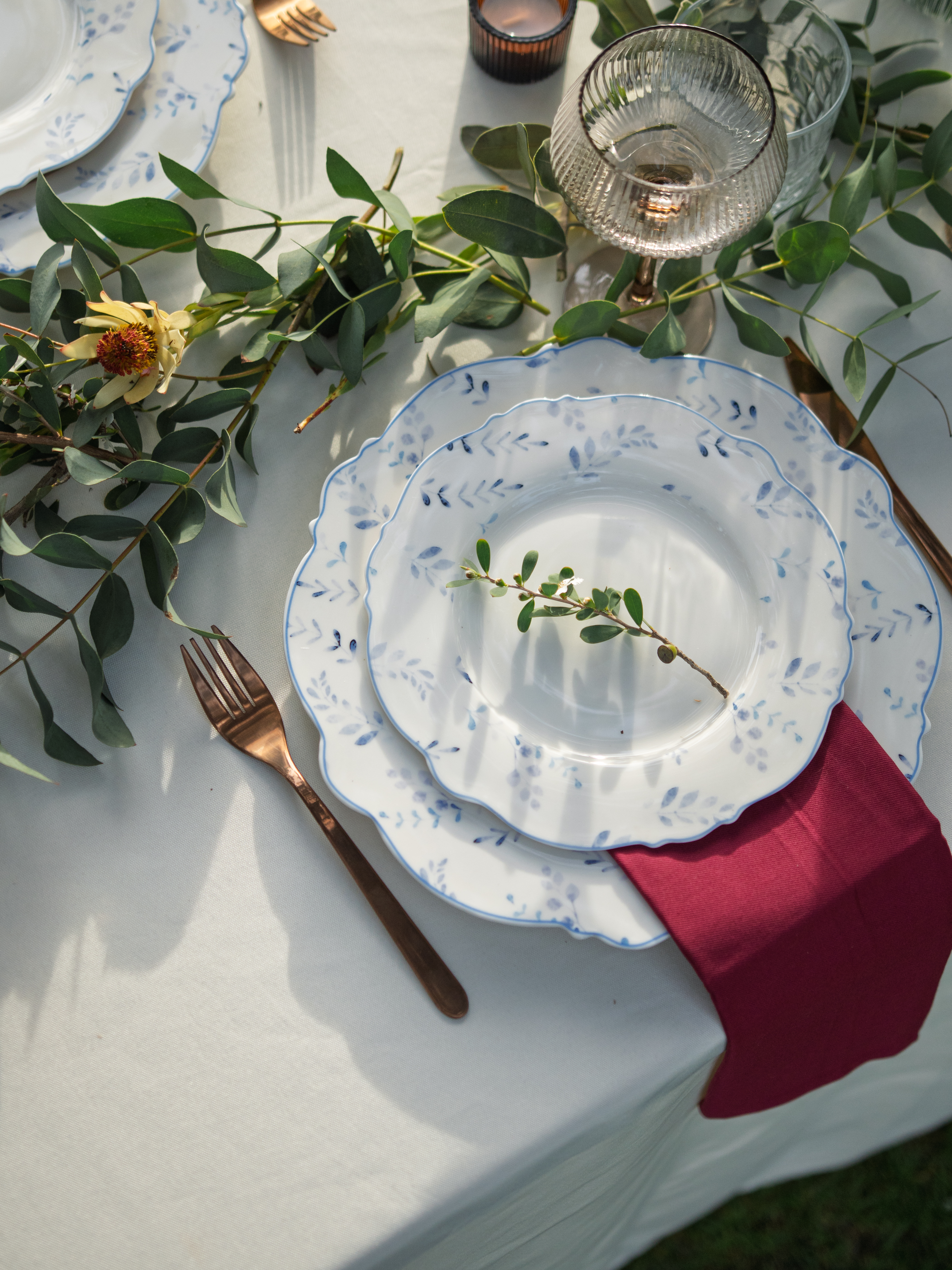 Elegant place setting with blue and white china, burgundy napkin, rose gold flatware, and eucalyptus — table styling by KarolinaBLevents